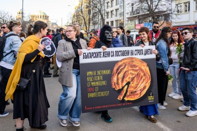 A crowd of demonstrators are gathered on the street in a city. Three people, including a person in the center wearing a gorilla mask, hold up a large sign with an image of a traditional Bulgarian pastry with a tiny slice cut out of it and text in Bulgarian. 