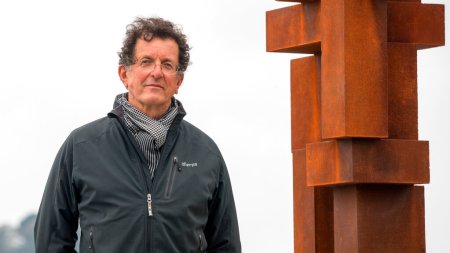 A man stands next to an abstract sculpture made of what appears to be weathered steel.